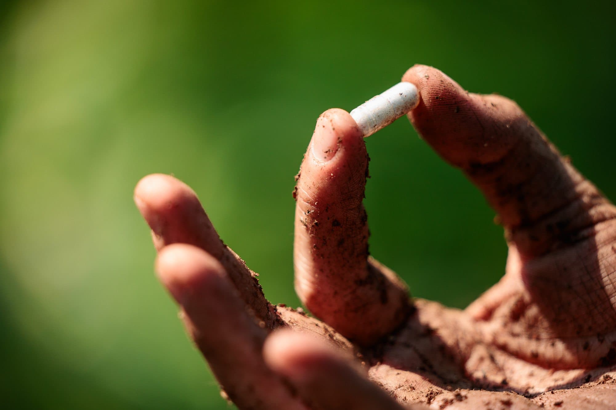 A soil-covered hand holding up a single microbial capsule between two fingers